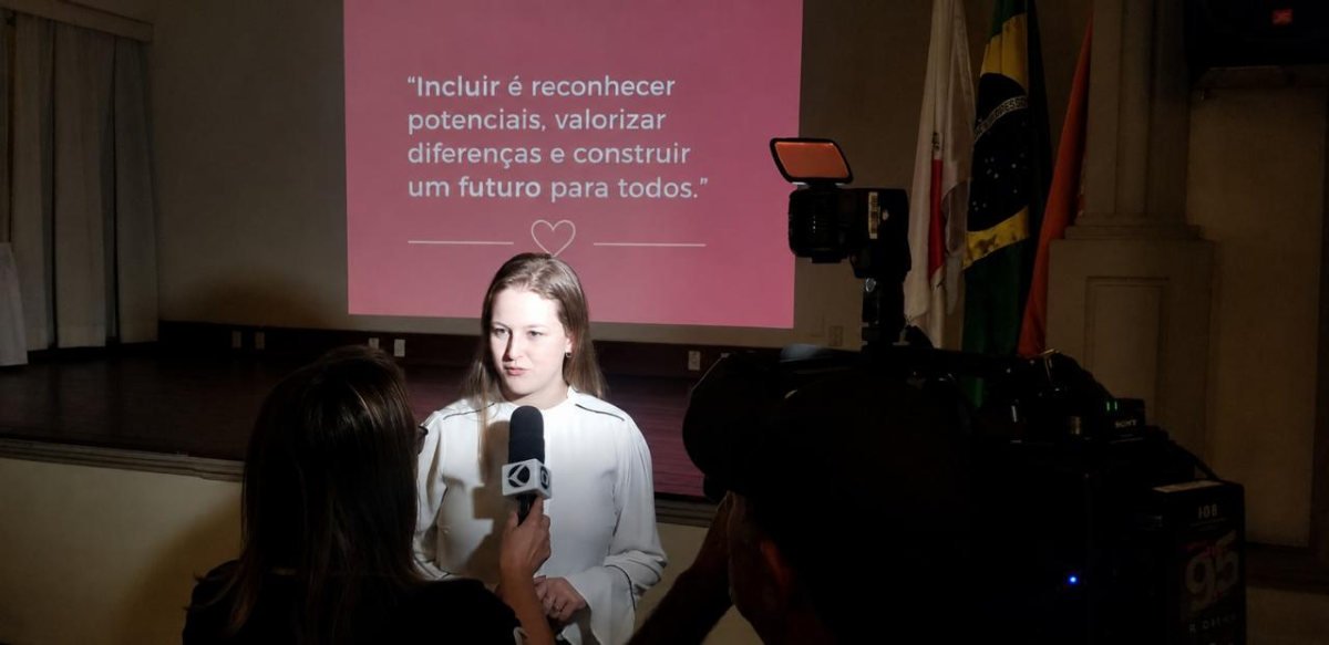Autora concedendo entrevista à TV durante o lançamento, diante do telão com a frase “Incluir é reconhecer potenciais”.