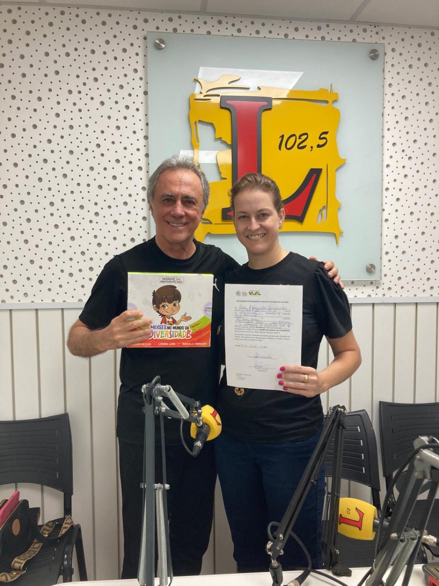 Lorena Lima no estúdio da Rádio Líder 102,5 segurando um livro da coleção, ao lado do apresentador.